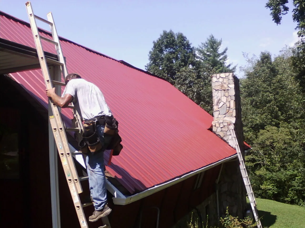 Roofer on a ladder beside a red metal roof and stone chimney — Soffit & Fascia Repair by Roofing Brothers in Porter